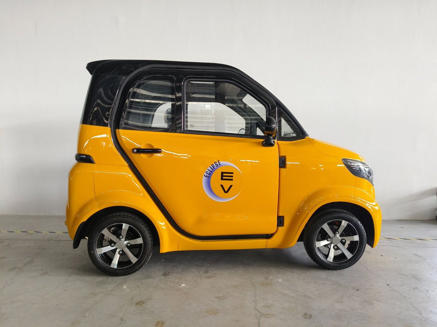 The Eclipse EV Nova, a compact and stylish yellow electric vehicle, provides comfortable transportation. Parked indoors against a plain white wall, the car features a black roof and windows, four doors, and an "EV" logo with a plug symbol. Its alloy wheels shine next to its powerful 60V 58Ah lead-acid battery.