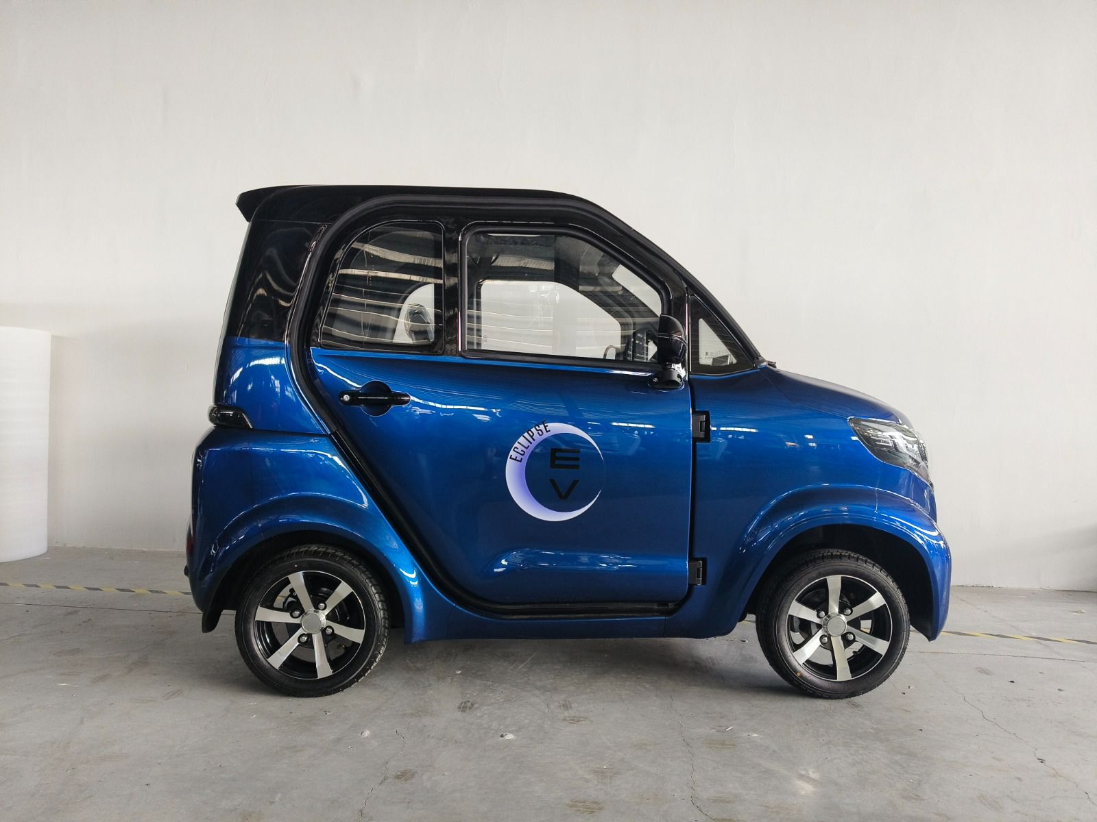 A compact, upright designed blue Eclipse EV Nova is parked indoors. It features a glossy finish, black accents, and an "EV" logo on the side. This electric vehicle sits on shiny alloy wheels and offers comfortable transportation powered by a 60V battery.