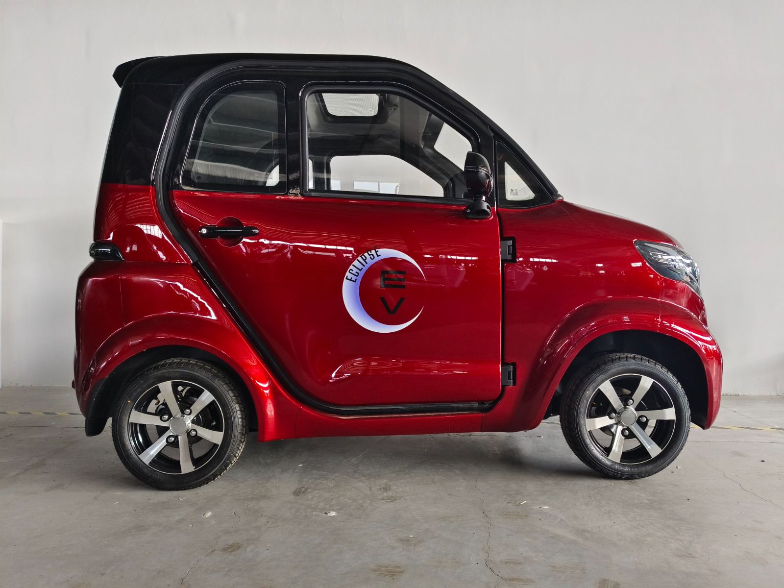 A red two-seater electric microcar, the Nova EV by Eclipse EV, is parked indoors against a plain wall. The car features a compact design with a black roof and five-spoke alloy wheels. Powered by a 60V 58Ah lead-acid battery, it promises comfortable transportation. The logo "Rewatt" with a stylized plug icon adorns the door.