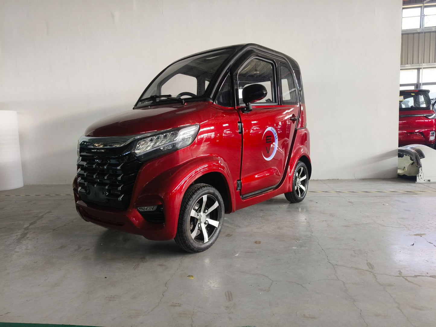 A modern red Eclipse EV Nova with black accents is parked indoors against a white wall. The car features a sleek, aerodynamic design with large windows and alloy wheels. A blue and white Eclipse EV logo is displayed on the side door.